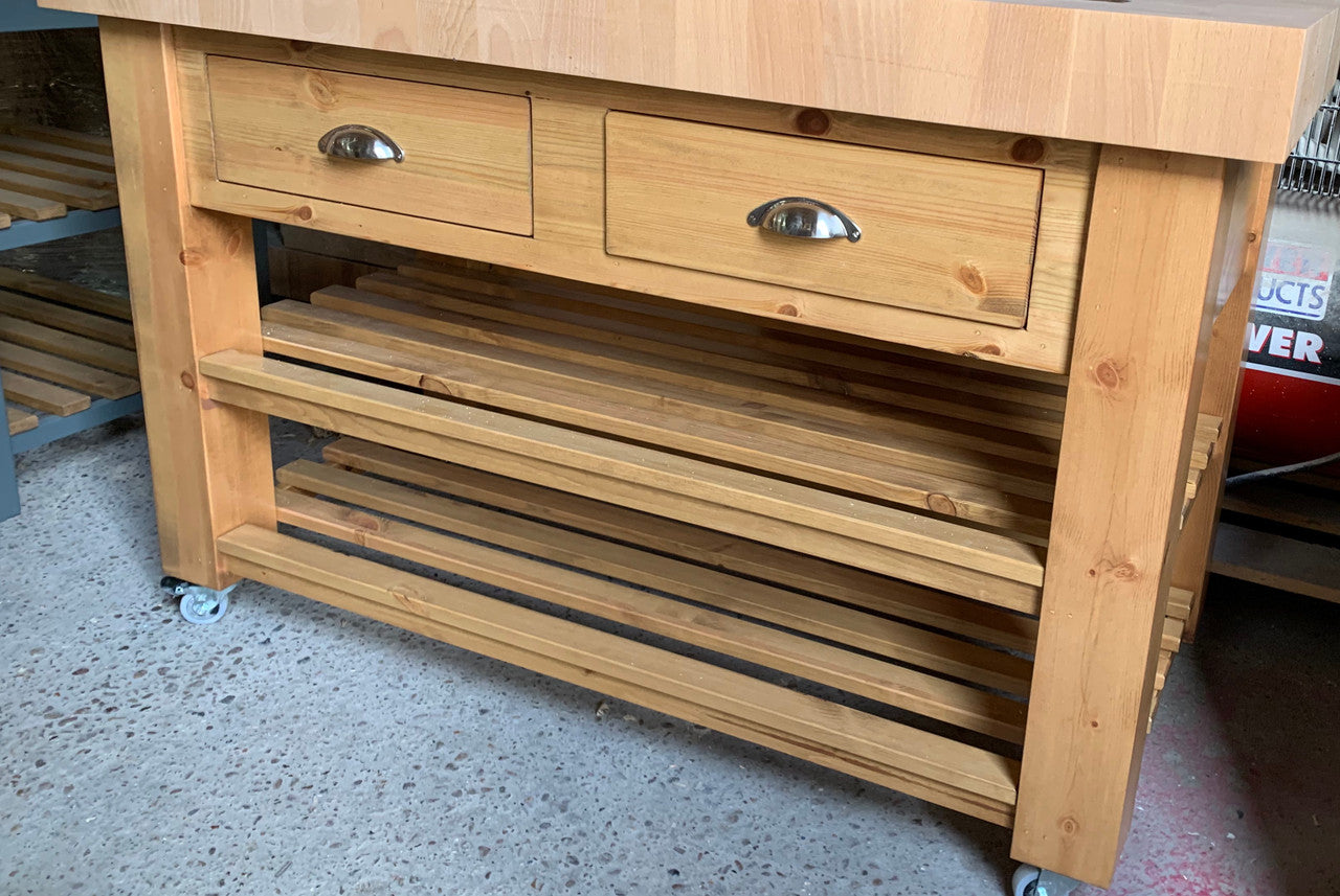 Wooden kitchen island with drawers and shelves on wheels in a storage area.